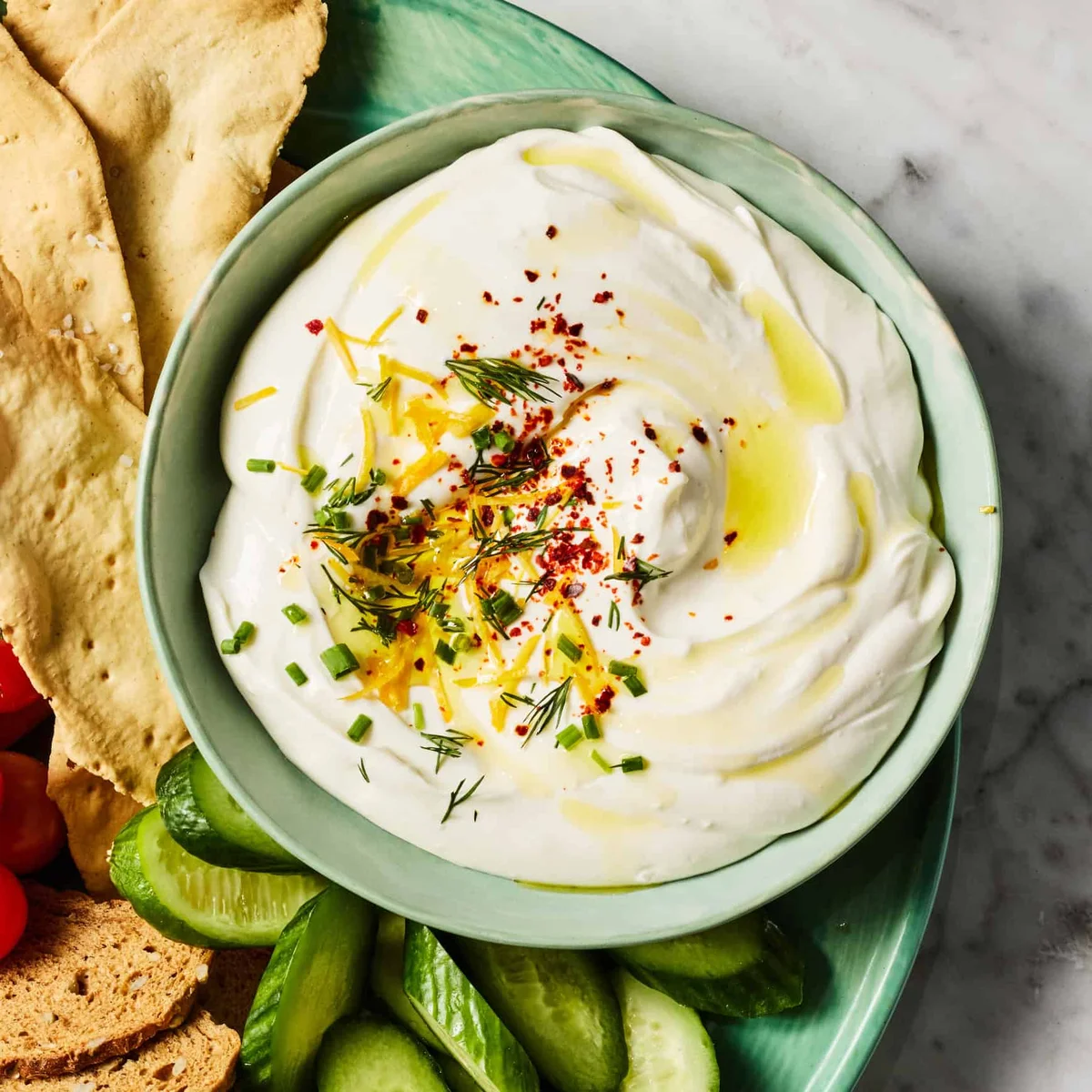 Whipped cottage cheese dip garnished with herbs, lemon zest, and olive oil on a two-tier platter with vegetables and pita