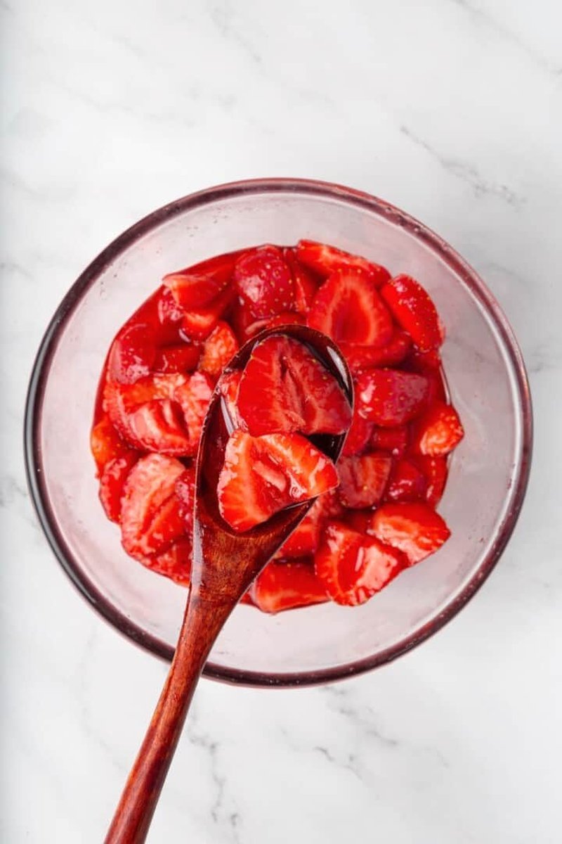 Spoonful of syrupy macerated strawberries being lifted from a glass bowl
